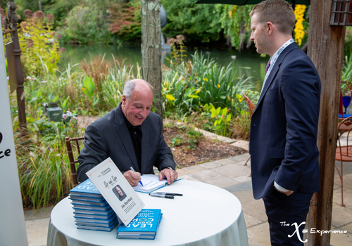 Jay Steinfeld at a table, signing his book for another attendee while he looks on
